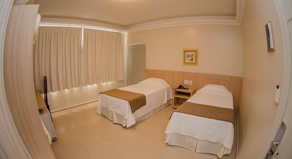 Quarto de hotel com duas camas, uma mesa de cabeceira com abajur, uma janela com cortinas e um quadro na parede.