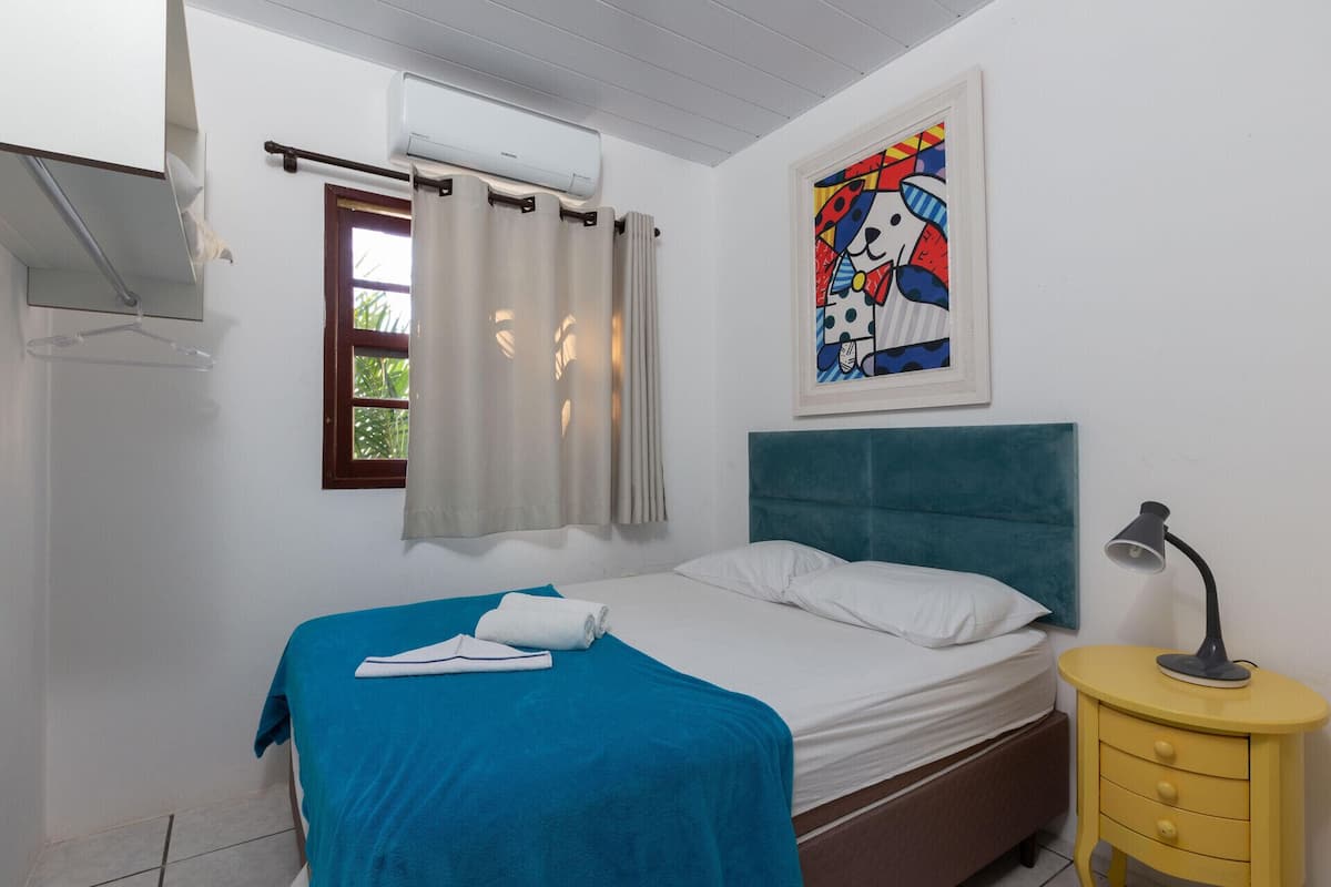 Une chambre à coucher avec un lit, une fenêtre avec des rideaux, un appareil de climatisation, un tableau et une table de chevet avec une lampe.