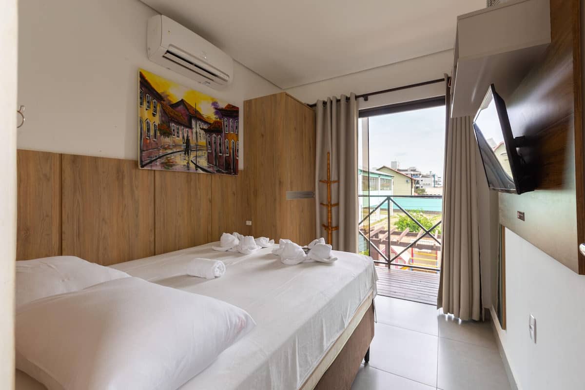 Une chambre d’hôtel avec un lit, un tableau, un climatiseur, une télévision et une vue sur un balcon donnant sur une piscine.