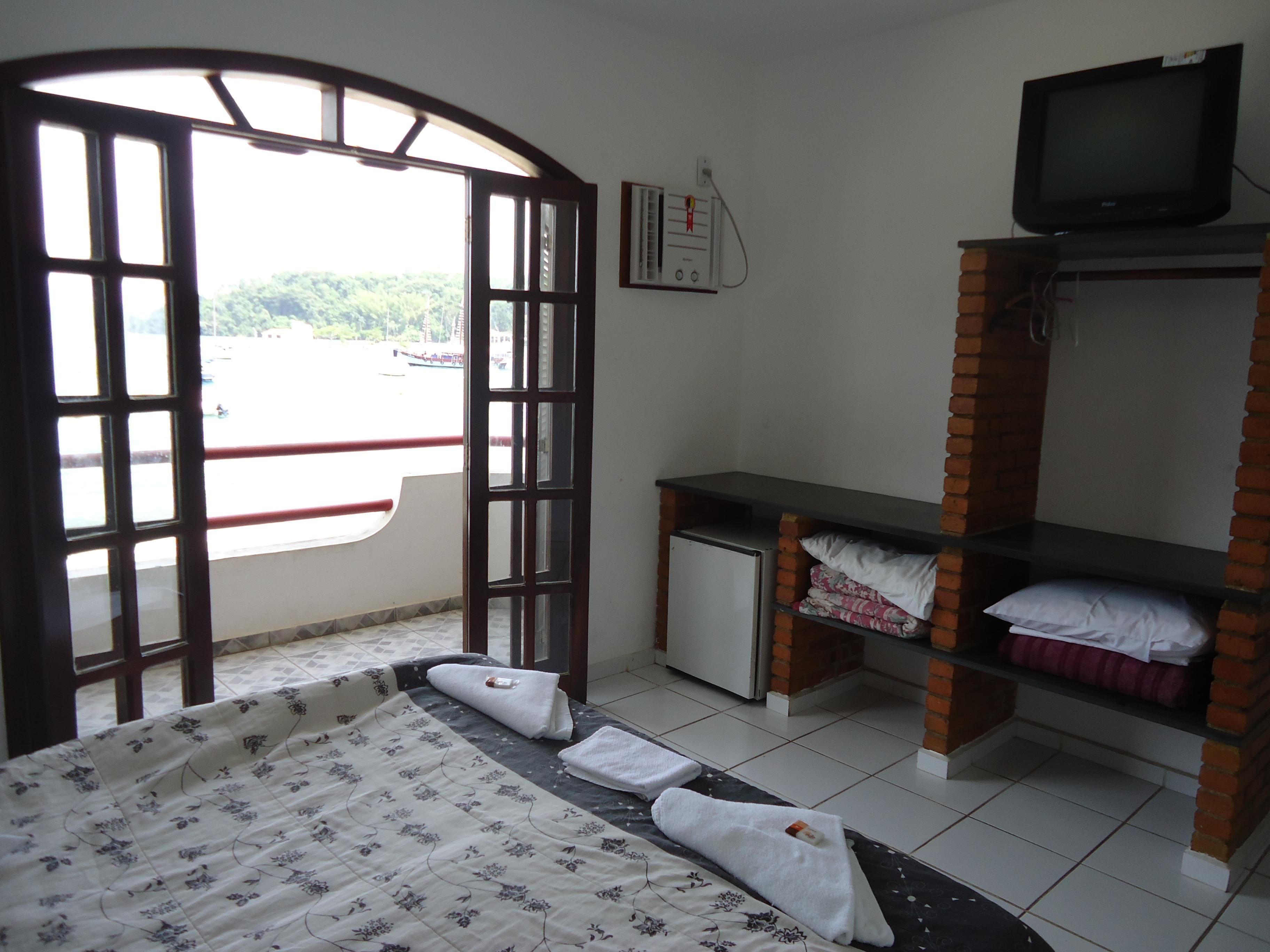 Quarto com uma cama, uma pequena escrivaninha com televisão, uma janela com vista para o verde e uma estante de tijolos.