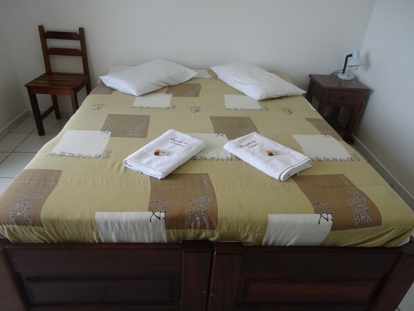 A neatly made bed with two white towels, a wooden chair, a bedside table with a lamp, and a wall with a light switch. - Pousada dos Corsários de Angra (Angra dos Reis)