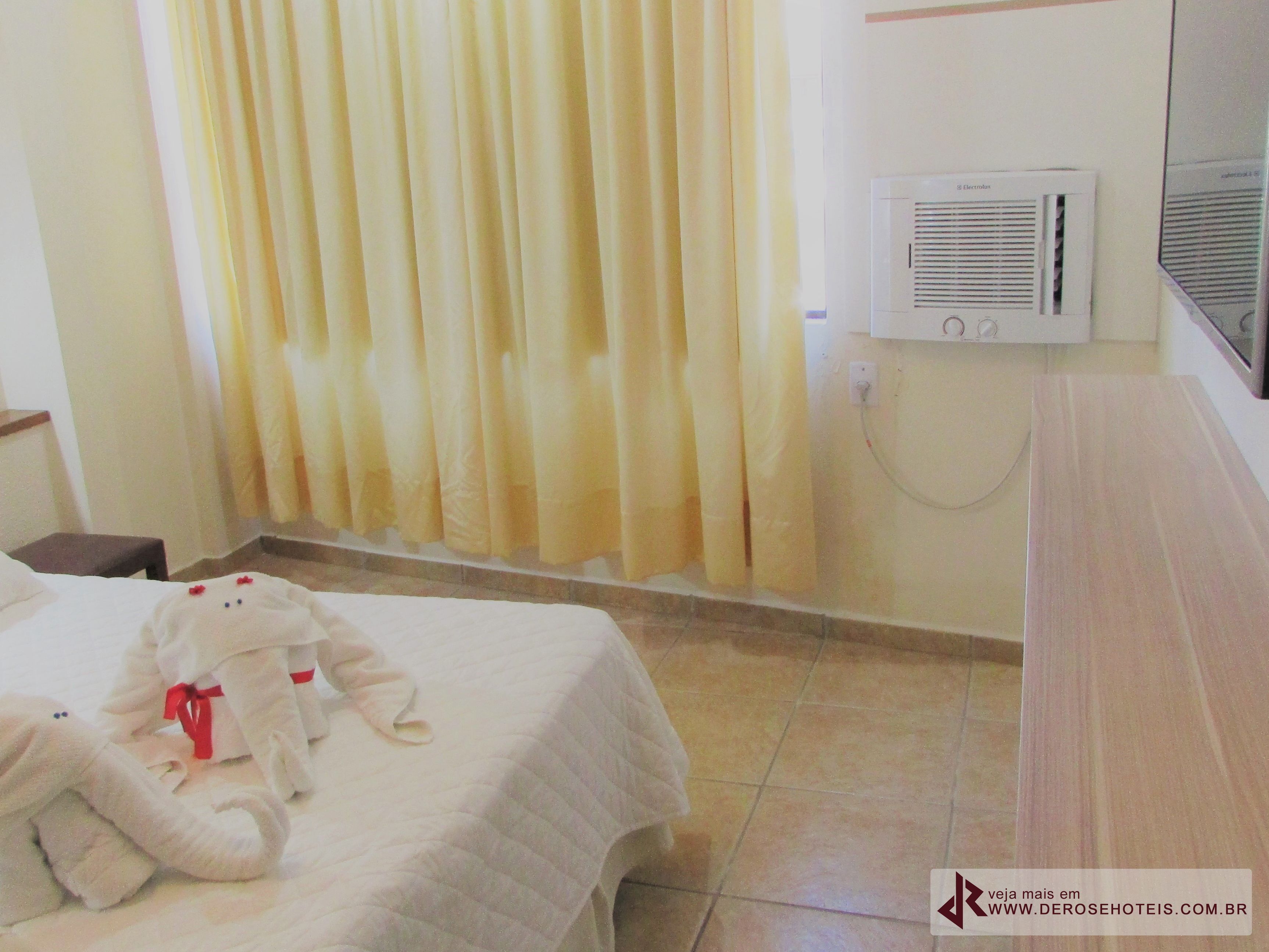 A hotel room with a bed, a teddy bear, curtains, an air conditioning unit, and a tiled floor.