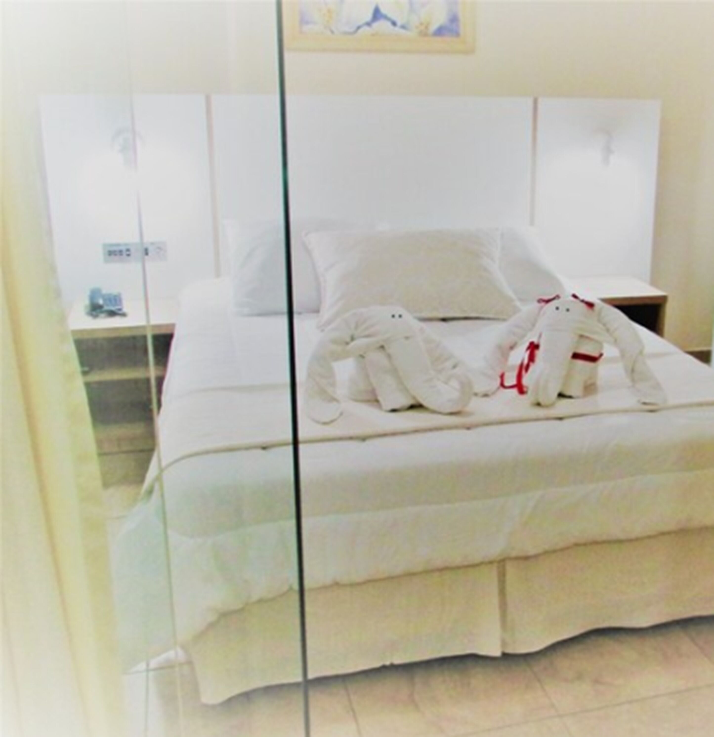 A neatly made bed with white linens and two plush white towels with red accents, a bedside table with a lamp, and a wall-mounted nightstand.