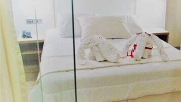 A neatly made bed with white linens and two plush white towels with red accents, a bedside table with a lamp, and a wall-mounted nightstand.