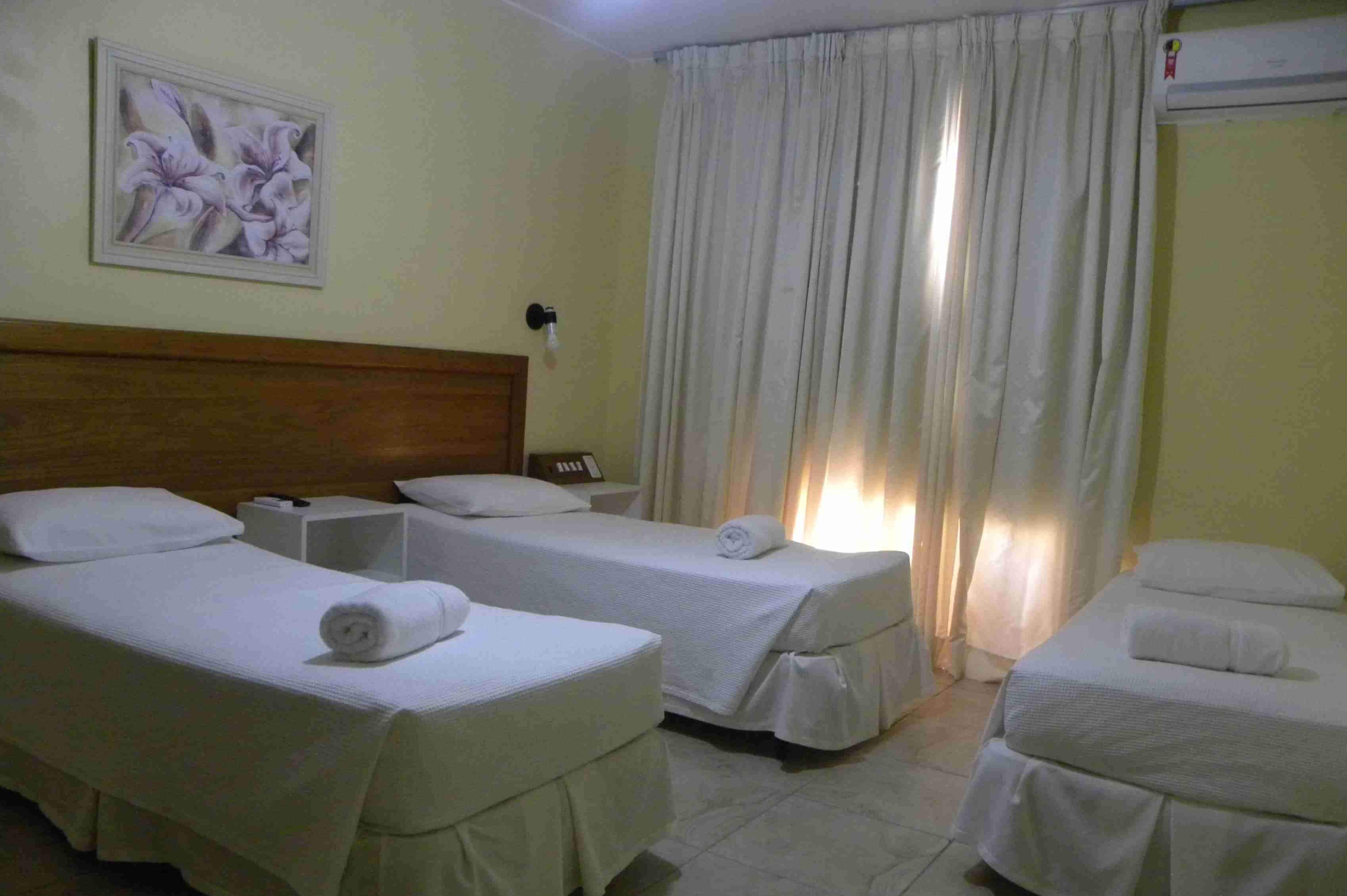 A hotel room with two single beds, a wooden headboard, a nightstand, a lamp, and a painting of flowers on the wall.