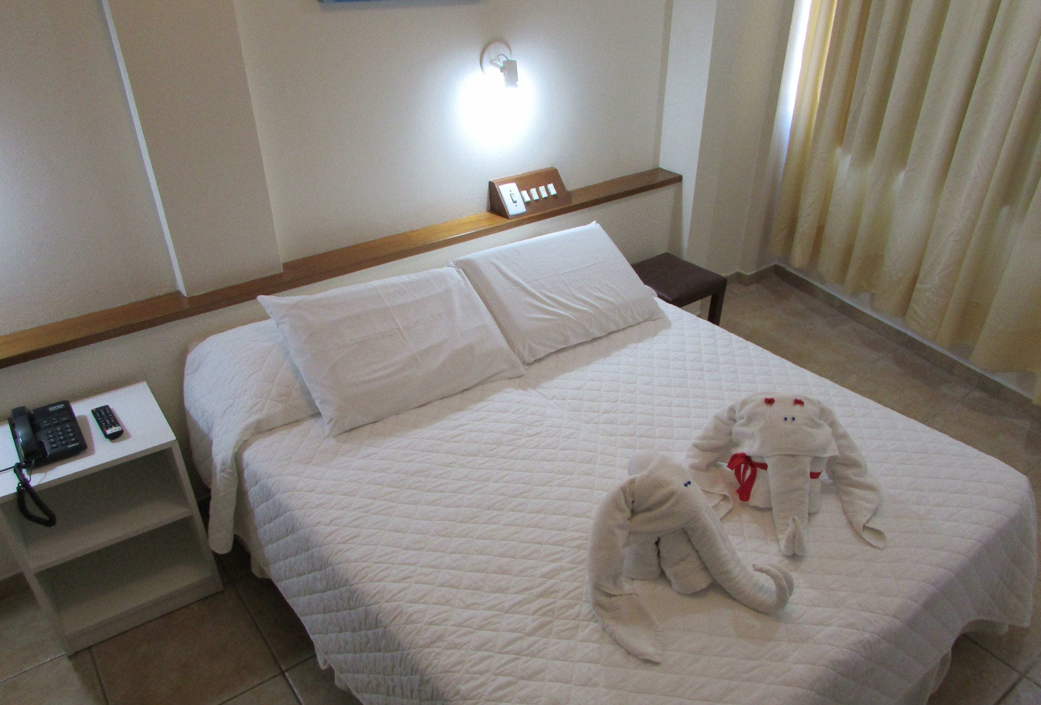Cama bem arrumada com manta acolchoada, mesa de cabeceira com telefone e luminária fixada na parede.