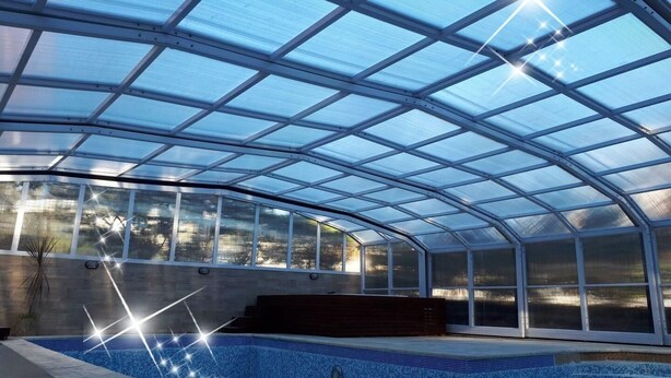 A covered swimming pool with a glass roof, surrounded by a concrete wall and a wooden deck. - Rey del Bosque (Valeria del Mar)