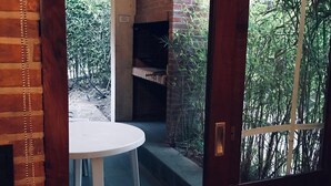 A patio area with a brick fireplace, a white table, and a view of a garden with bamboo and pink flowers. - Rey del Bosque (Valeria del Mar)