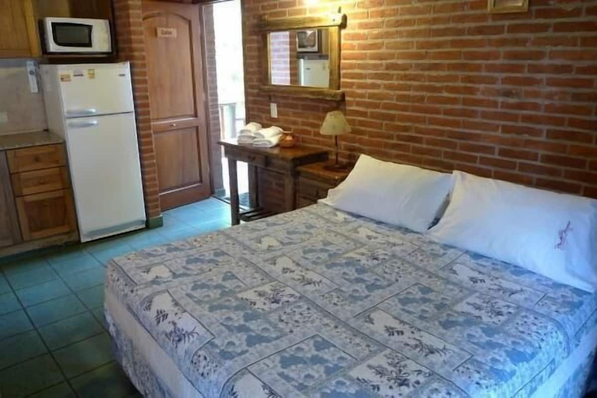 A bedroom with a patterned bedspread, a small bedside table with a lamp, and a brick wall.