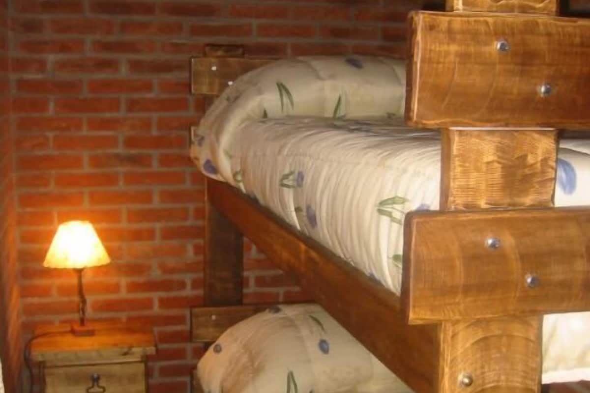 A bunk bed with a wooden frame and floral bedding, a nightstand with a lamp, and a brick wall background.