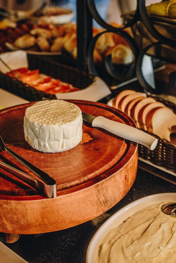 A cheese wheel on a wooden board with a cheese knife, surrounded by various food items. - Obba Coema Village Hotel (Capitólio)
