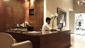 A reception desk with a computer, a telephone, and a vase with flowers. There is a framed picture on the wall and a window with a view of a car. - Hotel Marconi (Resistencia)