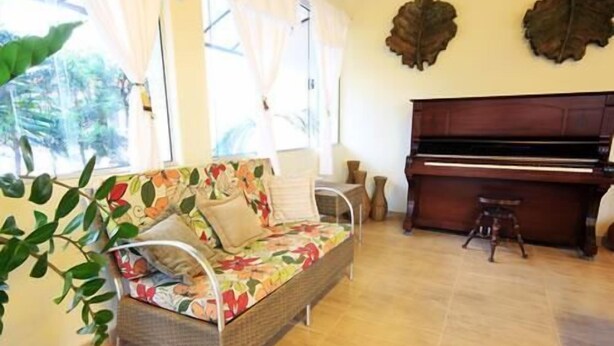 A room with a wicker sofa, a piano, and a large window with curtains. - Hotel Flamboyant (Olímpia)