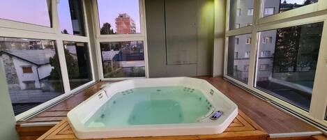 A hot tub on a wooden deck with large windows offering views of buildings and trees.