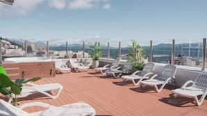 A rooftop terrace with white lounge chairs, potted plants, and a view of a city and mountains.