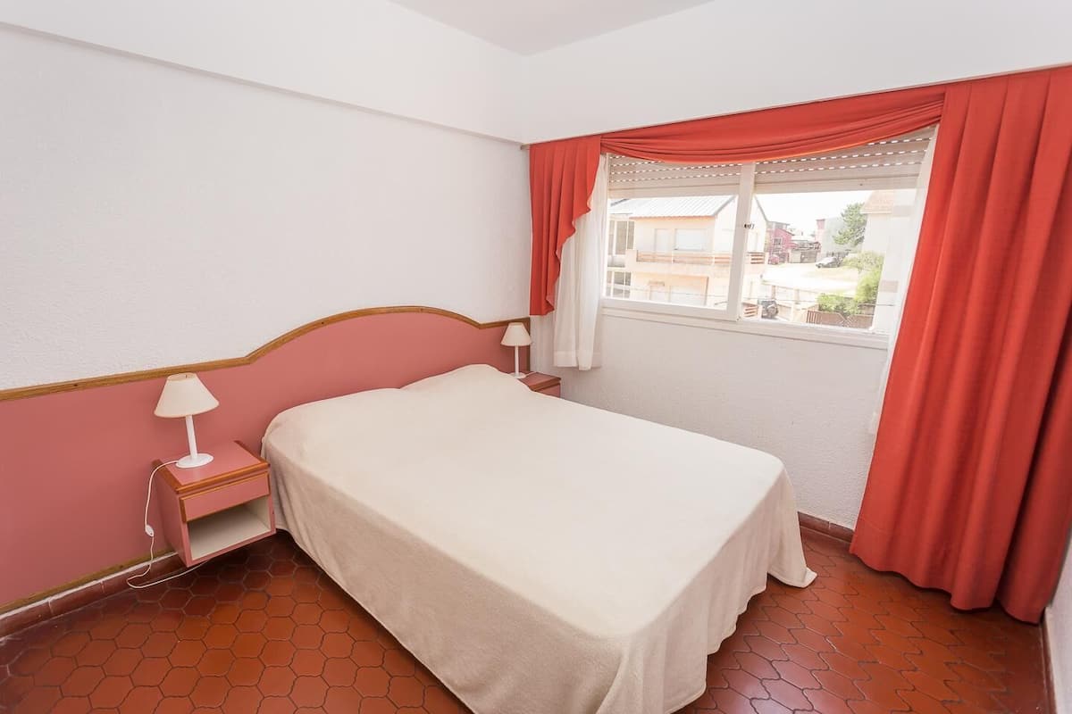 Un dormitorio con cama, mesita de noche, lámpara, ventana con cortinas y vistas a edificios.
