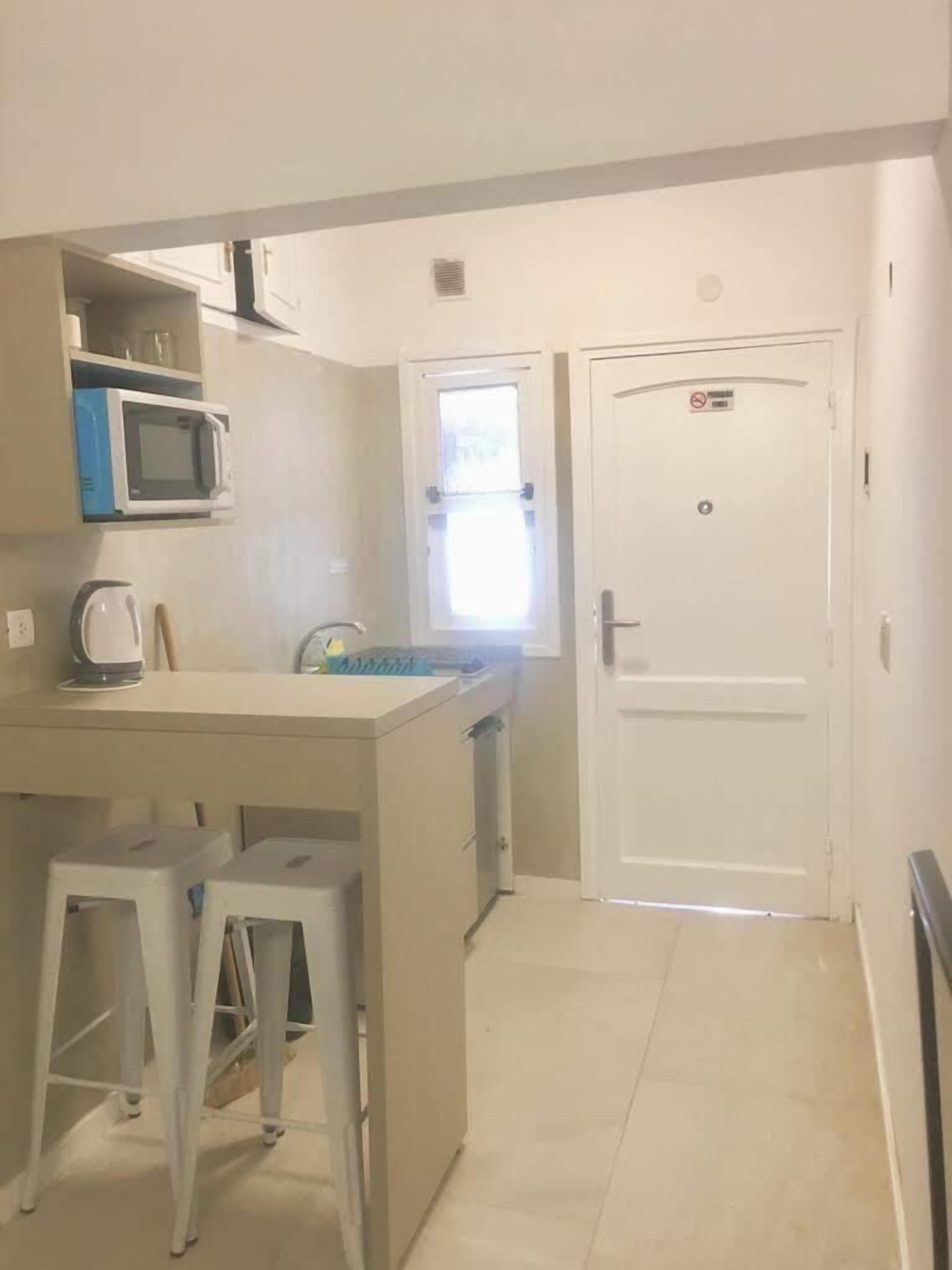 A compact kitchen with a microwave, a kettle, and a small island with stools. There is a door with a no smoking sign and a window with blinds.