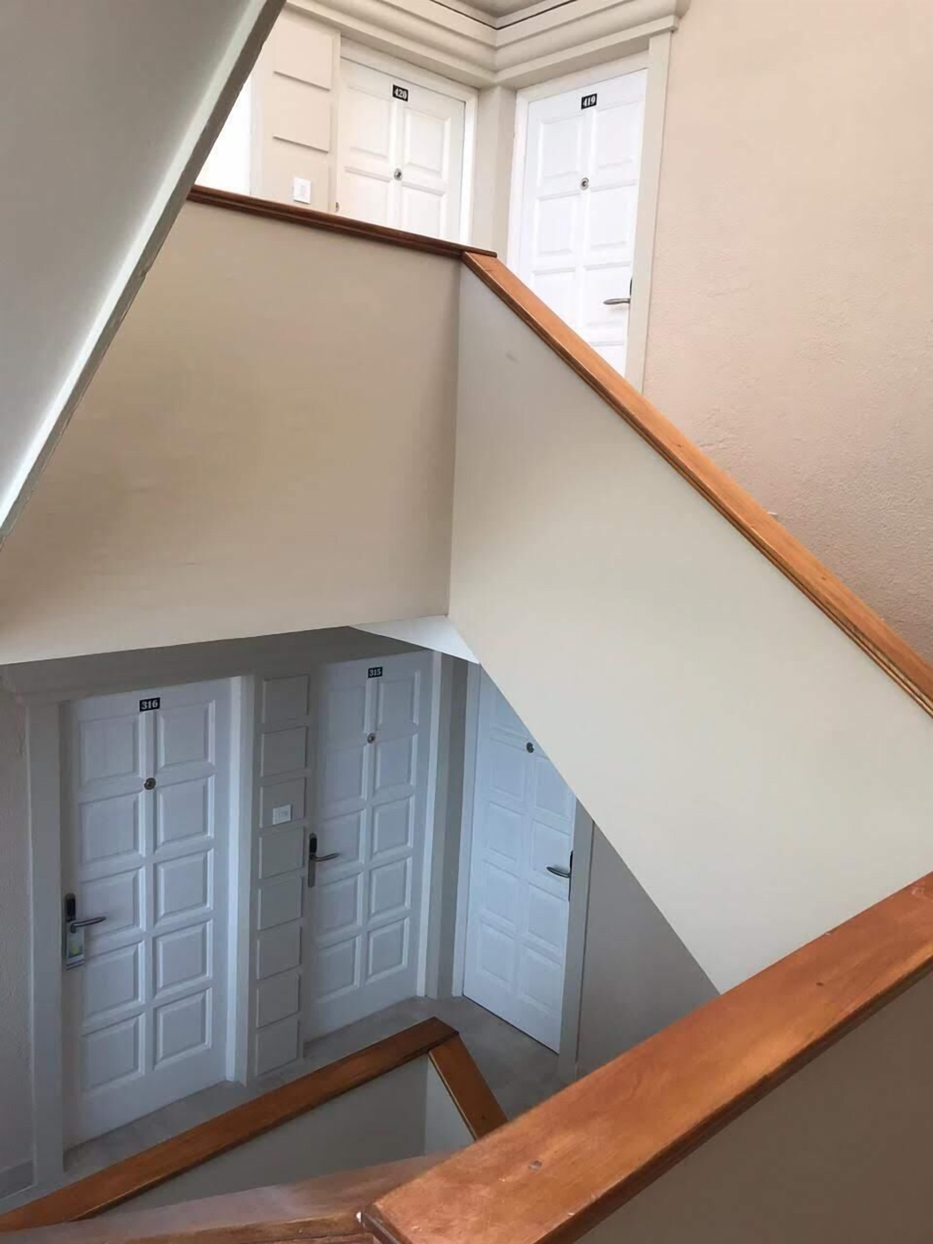 A multi-level interior with a wooden staircase leading to a landing. There are white doors with numbers on them, indicating room assignments.