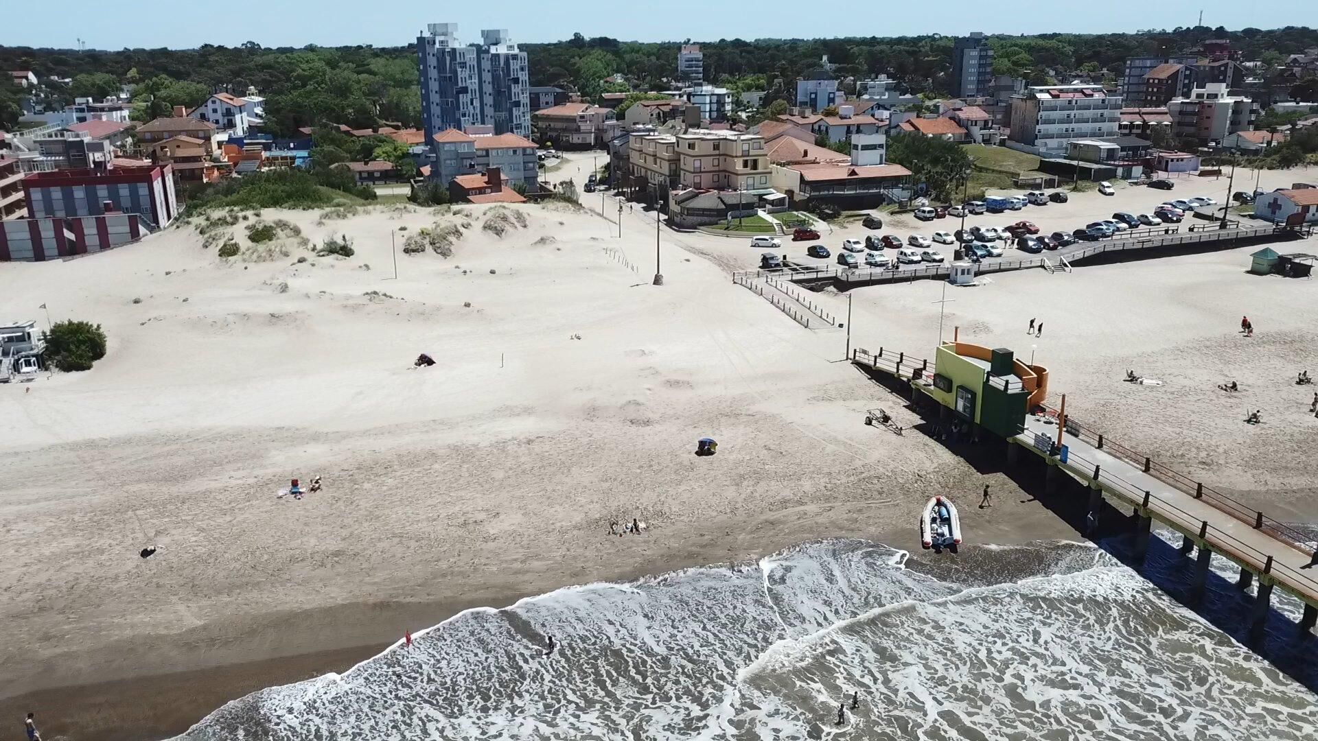 An aerial view of a beach with a pier, buildings in the background, and a few people on the sand.