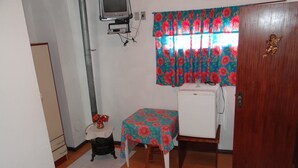 Una stanza modesta con pavimento in legno, un frigorifero bianco, un tavolo con tovaglia a fiori, un piccolo fornello nero e un televisore fissato alla parete.