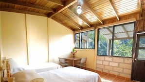 A room with two beds, a table with chairs, a lamp, and large windows with a view of greenery. - Hotel Panacam Lodge (Santa Cruz de Yojoa)