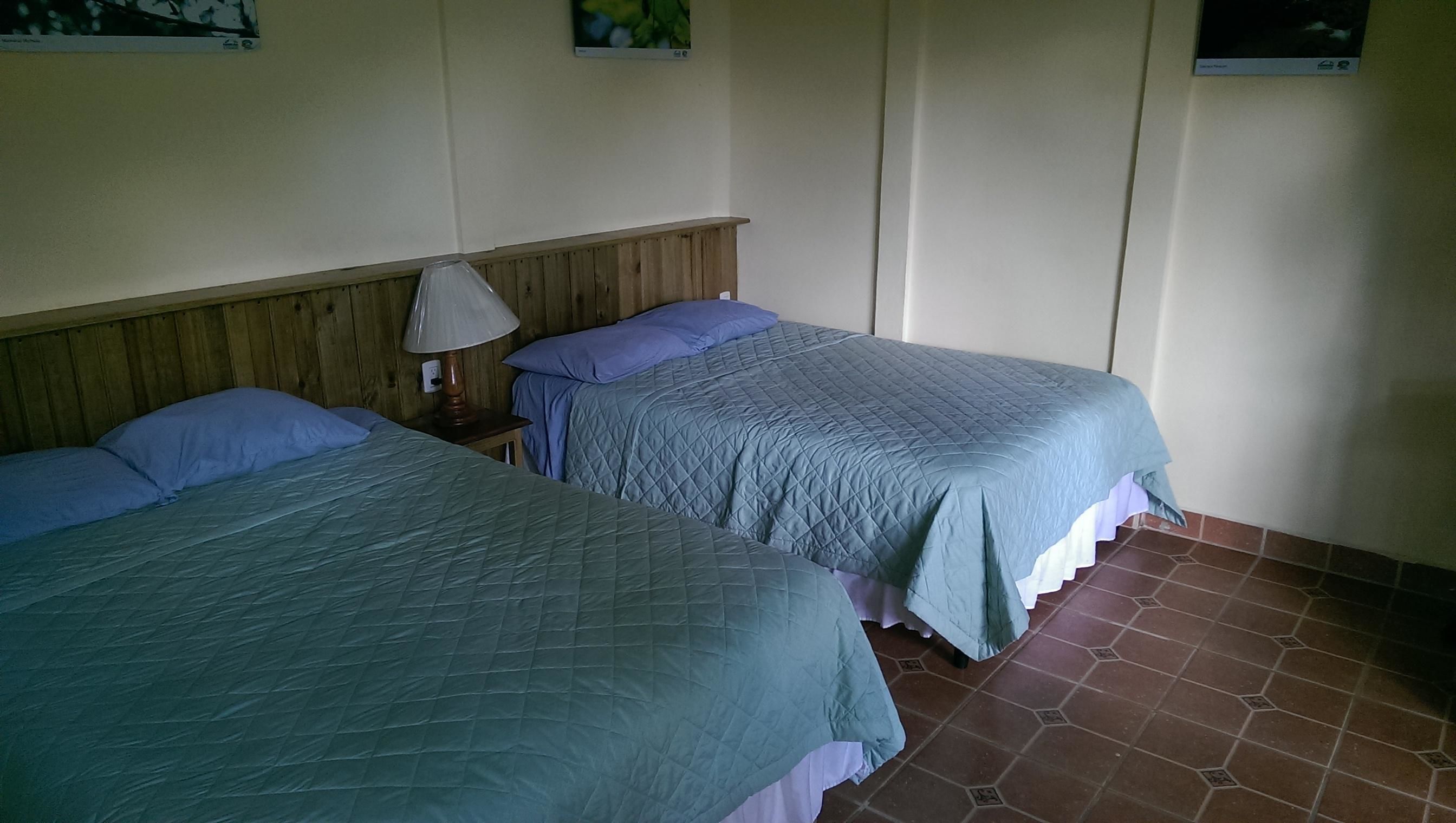 Two single beds with blue bedding, a wooden headboard, a nightstand with a lamp, and a tiled floor.