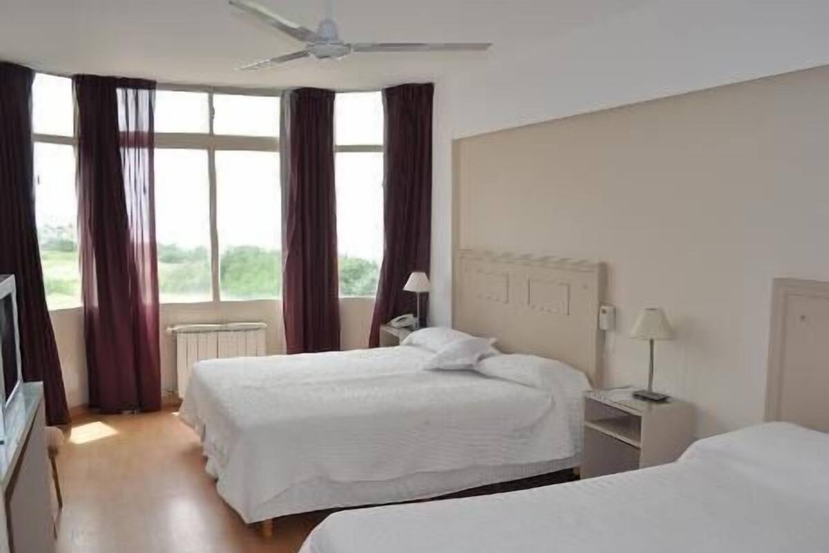 A hotel room with two beds, a ceiling fan, a window with curtains, a bedside table with a lamp, and a radiator.