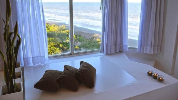 A room with a large window overlooking a beach, a white cushioned bench, and a potted plant.