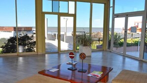 A spacious indoor area with large windows offering a view of the beach and ocean. There are wooden tables and chairs, and a potted plant.