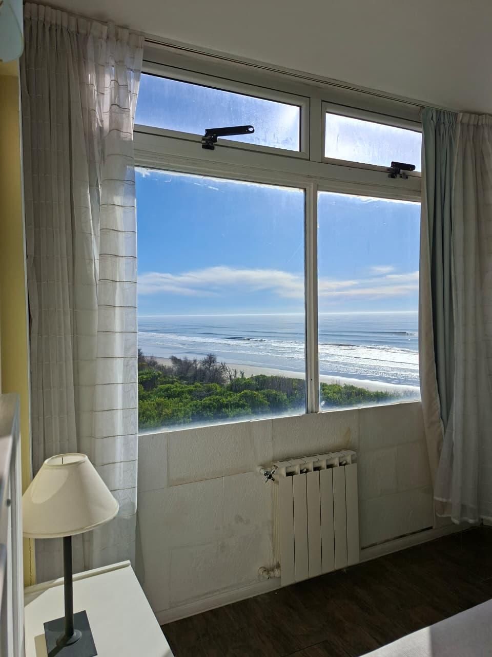 Una habitación con un ventanal grande con vista a una playa, un radiador debajo y una mesa con una lámpara.