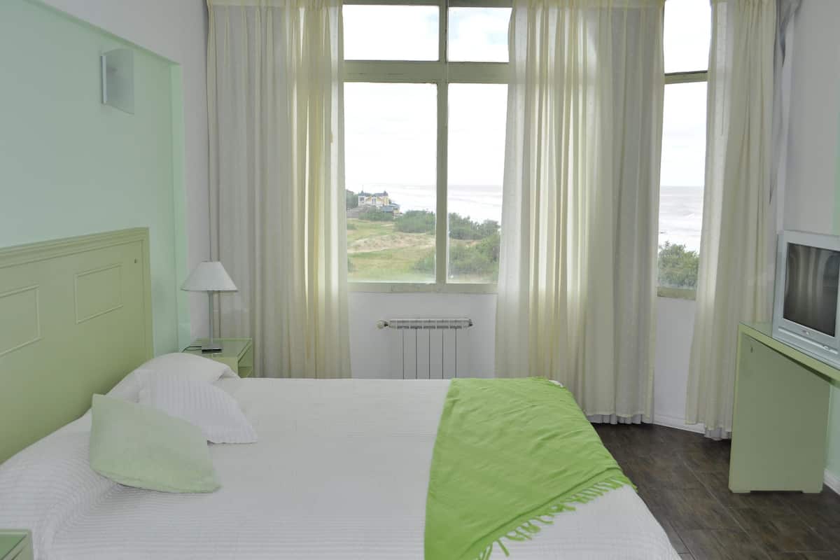 A hotel room with a bed, a TV, a window with curtains, and a view of the sea.
