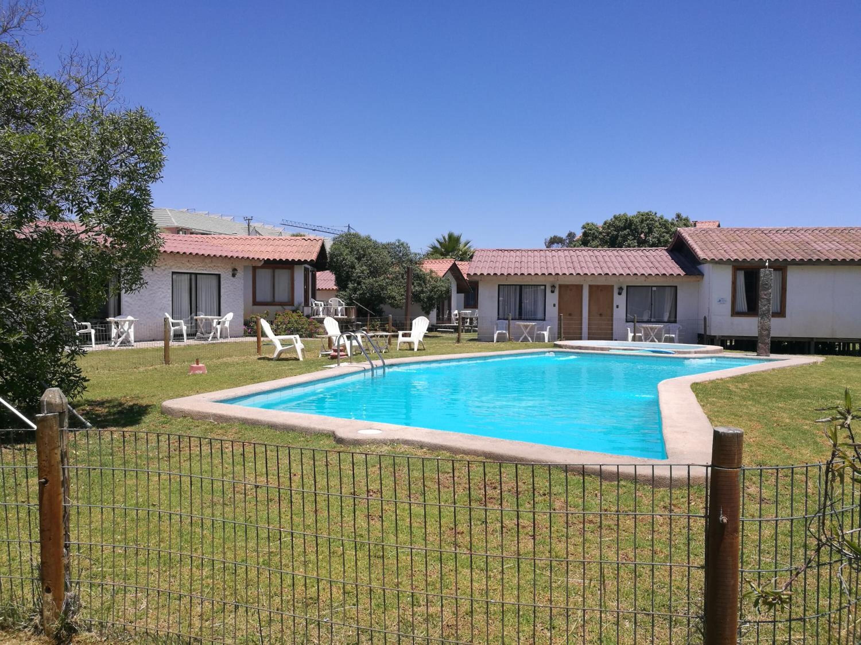 Zona de piscina con piscina cercada, sillas de lounge blancas y un edificio con tejado de tejas rojas.
