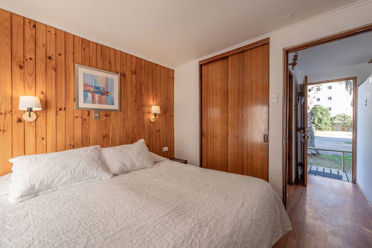 A bedroom with a wooden wall, a bed with white bedding, two wall-mounted lamps, and a door leading to an outdoor area.