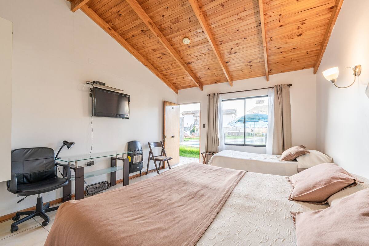 A hotel room with a wooden ceiling, a bed, a desk with a chair, a TV mounted on the wall, and a window with curtains.