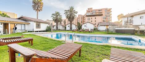 A poolside area with wooden loungers, a swimming pool, and residential buildings in the background.