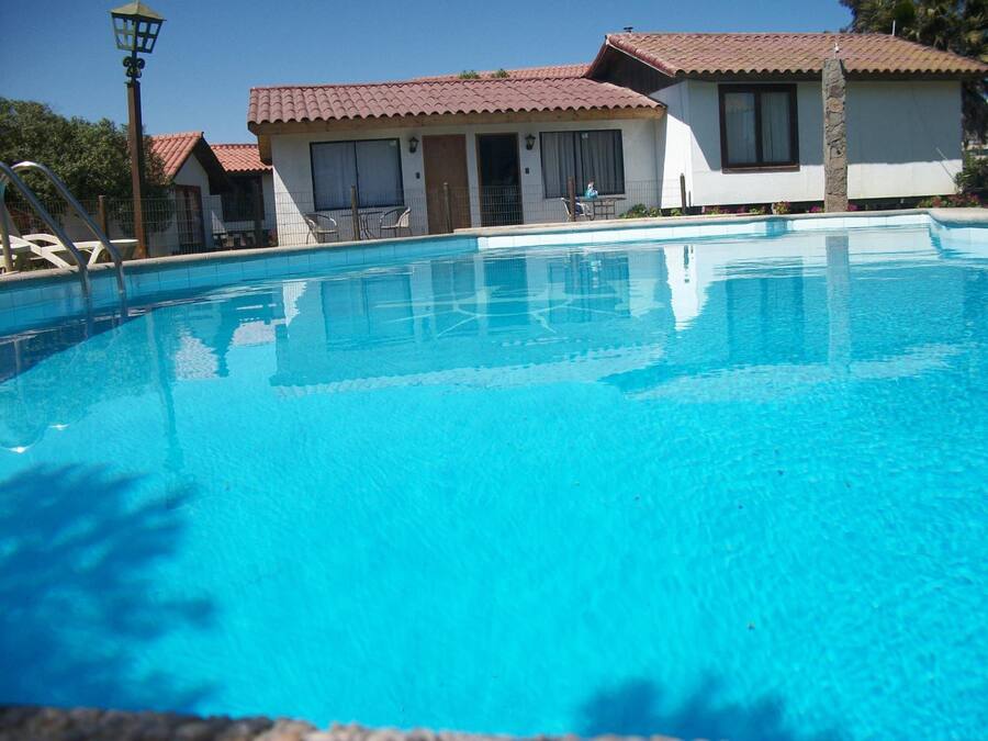 A clear blue swimming pool with a tiled deck, surrounded by buildings with red-tiled roofs and white walls.