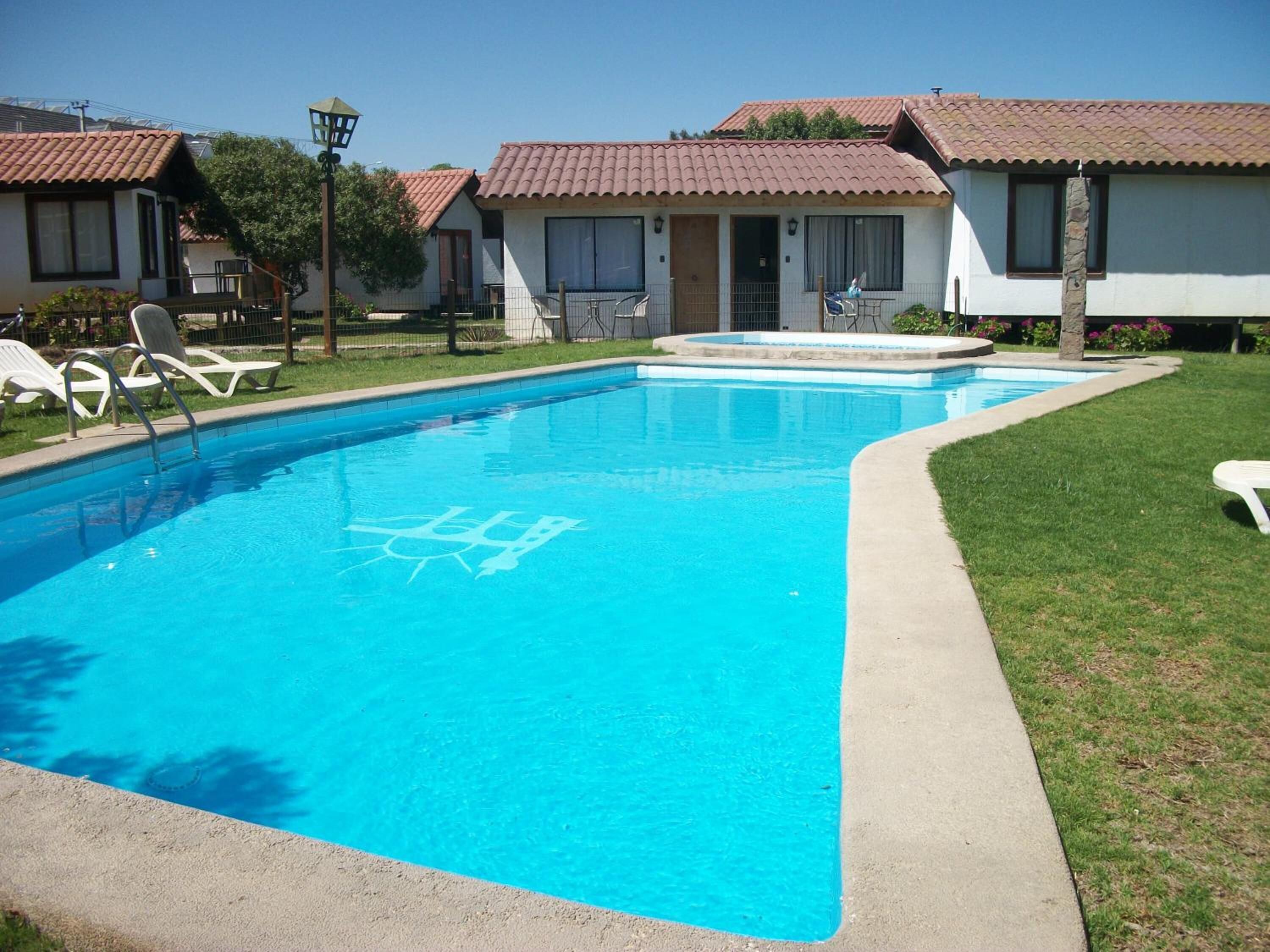 A swimming pool with a clear blue water, surrounded by a concrete deck and a grassy area. There are lounge chairs and a small building with a tiled roof nearby.