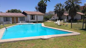 Una piscina con borde de baldosas, rodeada de césped y palmeras. Hay sillas de descanso blancas y una pequeña mesa cerca de la piscina.
