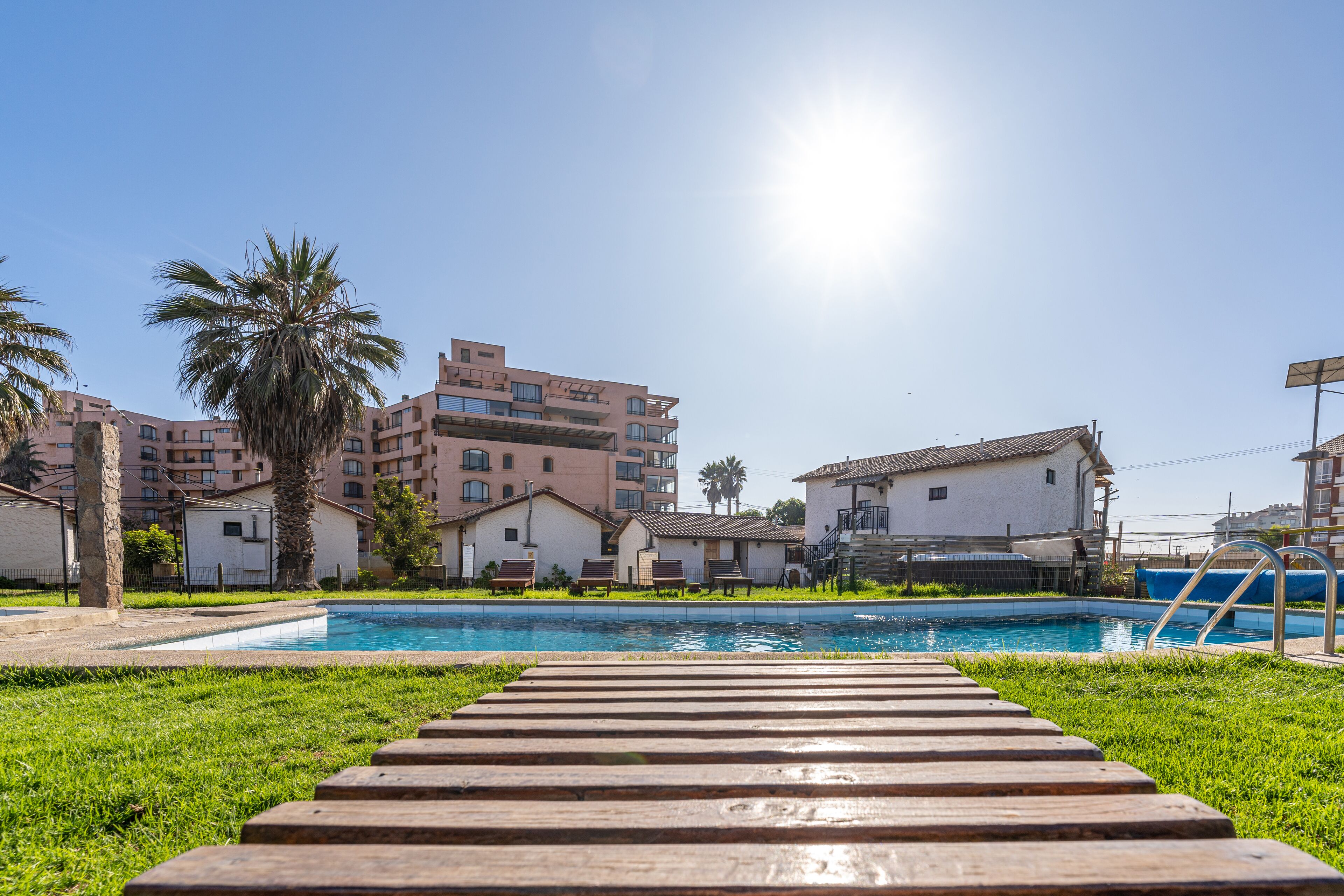 A swimming pool with a wooden deck leading to it, surrounded by greenery and buildings in the background.