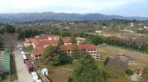 A residential area with a large building featuring multiple floors and a red-tiled roof. There is a fenced sports field in the foreground, and a road with parked vehicles is visible to the left. - Hotel Luz y Fuerza Villa Giardino (Villa Giardino)