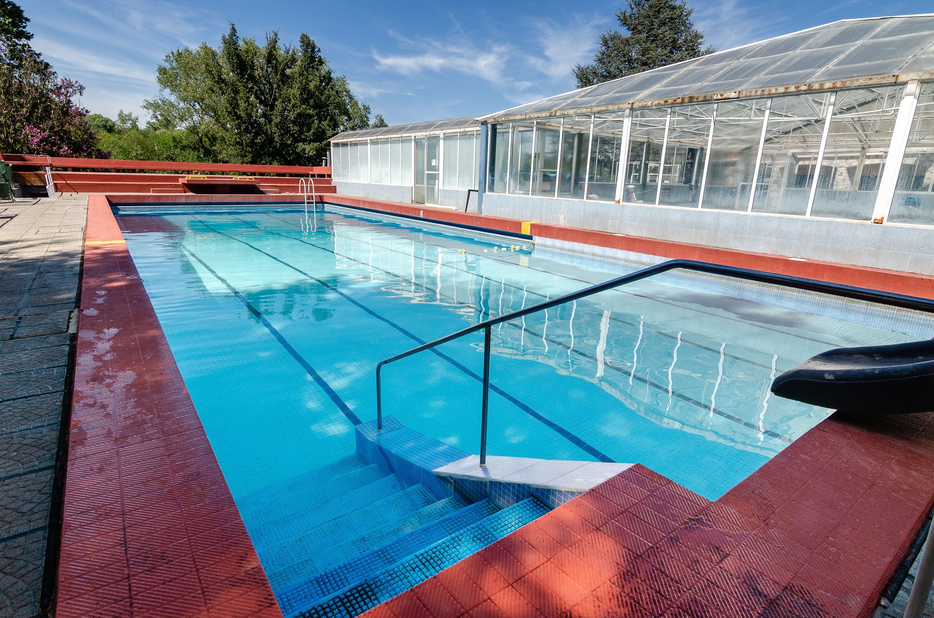 Ein Schwimmbecken mit Stufen, umgeben von einer roten Terrasse und einem Gewächshaus im Hintergrund.