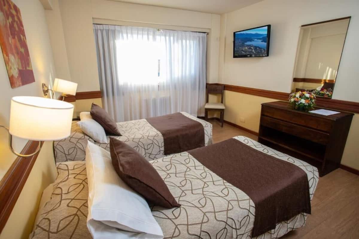A hotel room with two beds, a desk, a chair, a dresser, a TV, and a window with curtains.