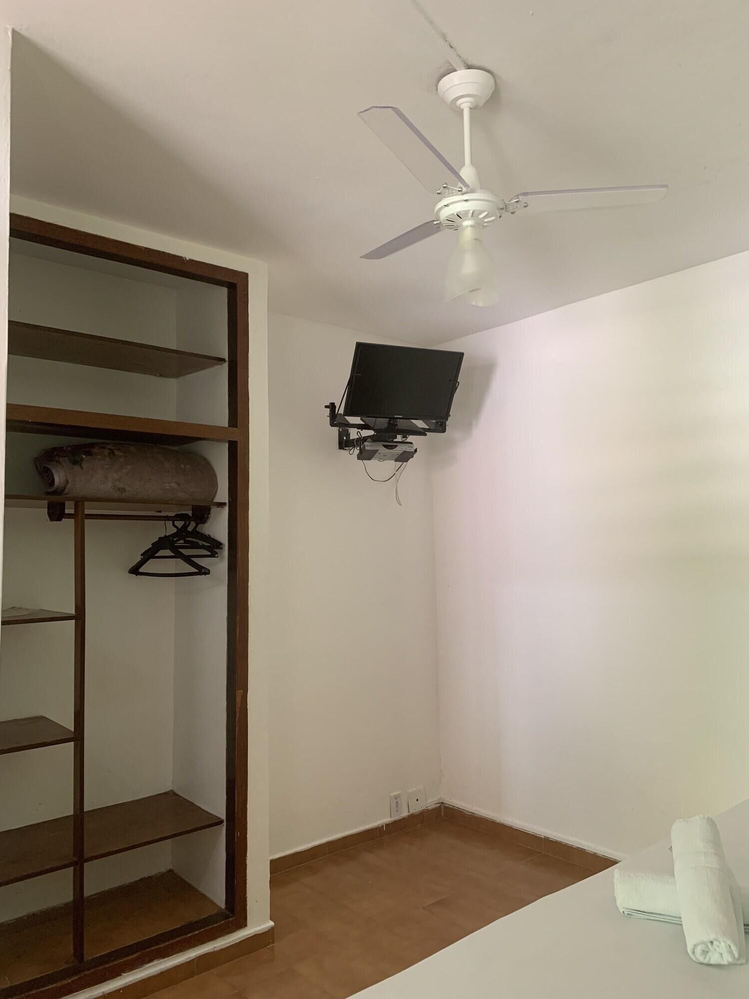 A room with a ceiling fan, a TV mounted on the wall, and a wooden wardrobe with shelves.