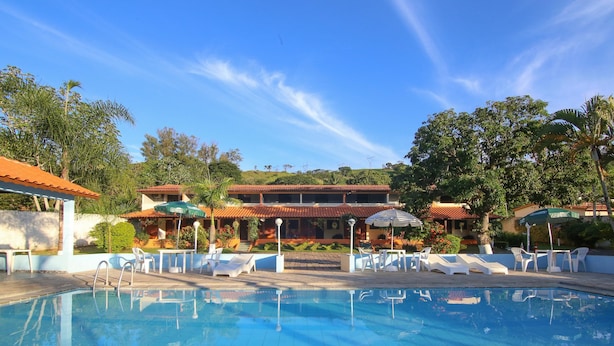 A pool area with lounge chairs and umbrellas, surrounded by buildings with red-tiled roofs and greenery. - Pousada Vale da Montanha (Serra Negra)
