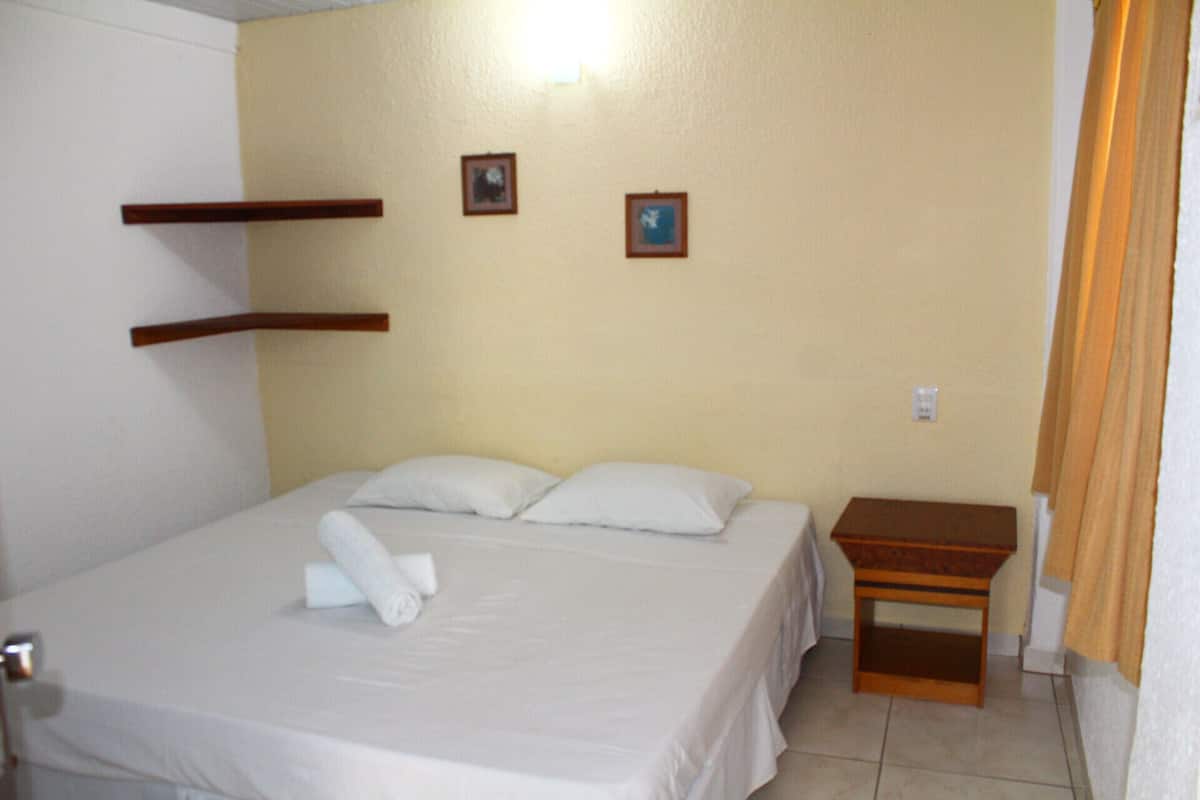 Cama bem arrumada com lençóis brancos e duas almofadas, mesa de cabeceira de madeira e dois quadros na parede.