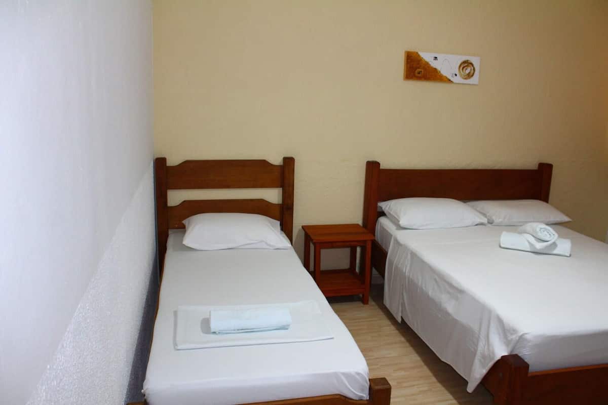Quarto com duas camas de solteiro, cada uma com cabeceira de madeira e roupa de cama branca. Há uma pequena mesa de cabeceira de madeira entre as camas, e um candeeiro fixado na parede acima da mesa.