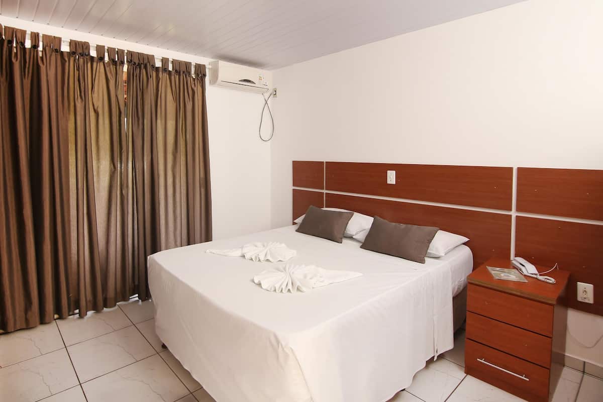 Quarto de hotel com cama bem arrumada, cabeceira de madeira, mesa de cabeceira com telefone e cortina.