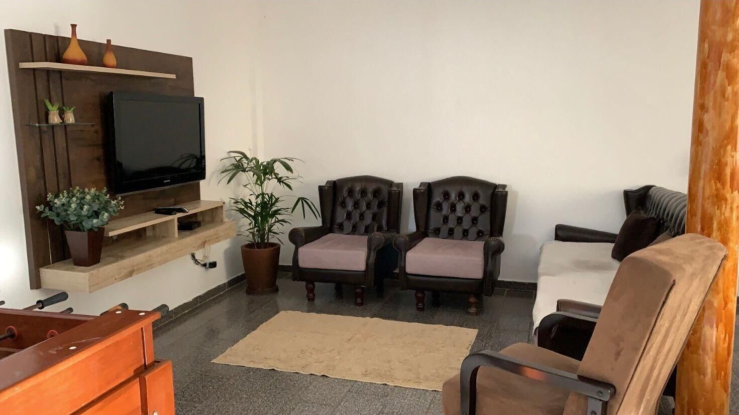 Uma sala de estar com televisão de tela plana, duas poltronas, um sofá, uma mesa de centro e uma estante de madeira com objetos decorativos.