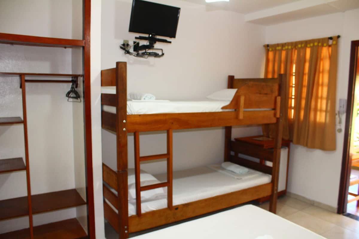 Quarto com beliche, televisão na parede, guarda-roupa de madeira e janela com cortinas.