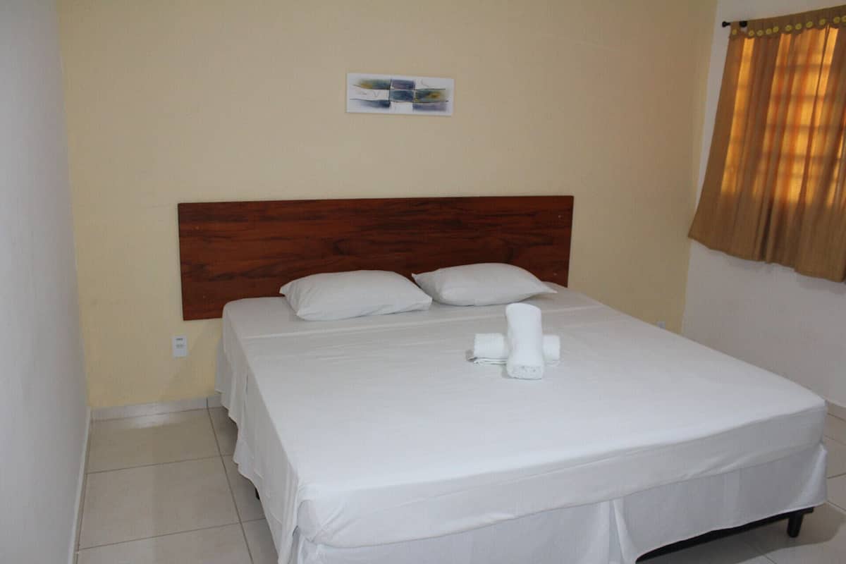 Uma cama bem arrumada com lençóis e almofadas brancas, uma cabeceira de madeira e uma pequena obra de arte na parede.